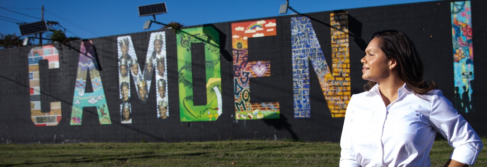 A Rowan student stands with her hand on her hip, looking right toward the sun with a mural behind her that paints the word Camden.