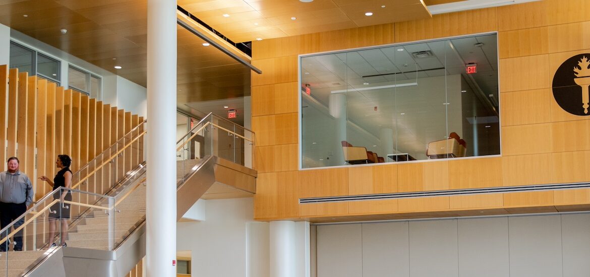 The wood panel interior of Business Hall at Rowan University, showing Rowan's torch emblem, a peekaboo cutout for a workspace at the top of the stairs, and two business majors talking while walking down the stairs.