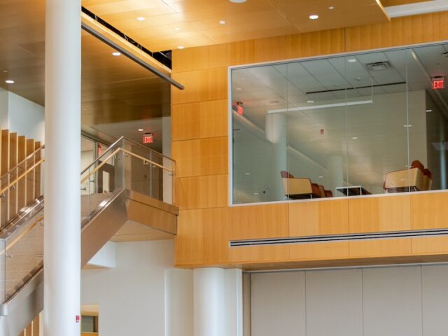 The wood panel interior of Business Hall at Rowan University, showing Rowan's torch emblem, a peekaboo cutout for a workspace at the top of the stairs, and two business majors talking while walking down the stairs.