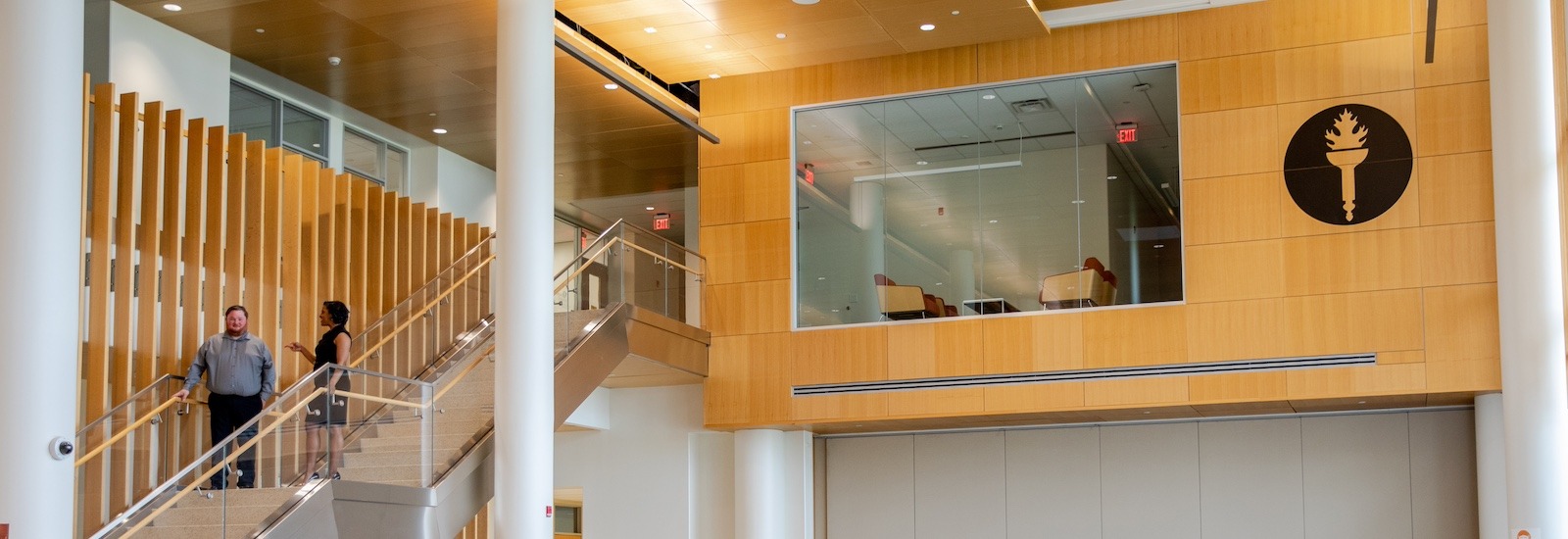 The wood panel interior of Business Hall at Rowan University, showing Rowan's torch emblem, a peekaboo cutout for a workspace at the top of the stairs, and two business majors talking while walking down the stairs.