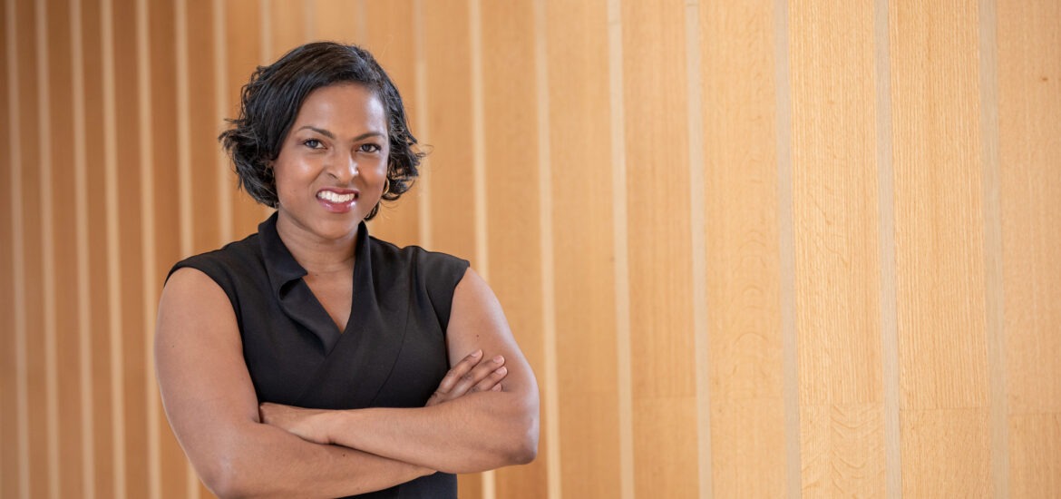 Female MBA candidate in a black sleeveless dress standing with arms crossed in front of a light wood-paneled wall.