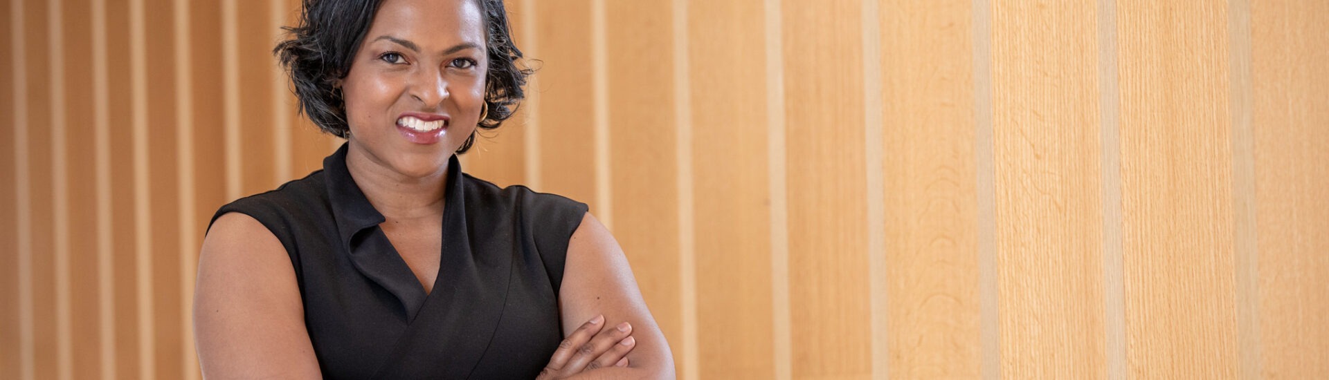 Female MBA candidate in a black sleeveless dress standing with arms crossed in front of a light wood-paneled wall.