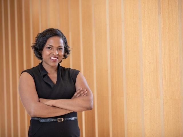 Female MBA candidate in a black sleeveless dress standing with arms crossed in front of a light wood-paneled wall.
