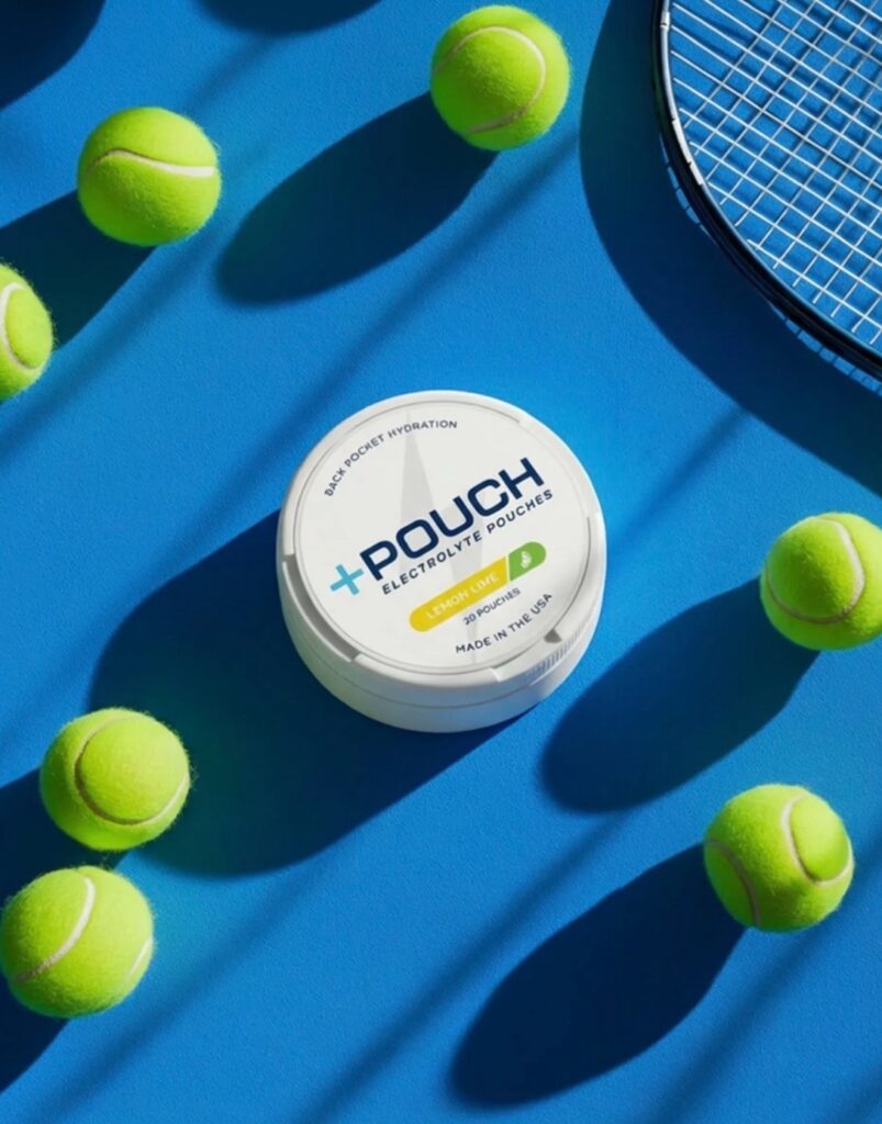 A professional product photo of a round, white metal tin that has the Plus Pouch logo in the center, on a blue background with dramatic shadows from tennis balls and a tennis racket around the product. 