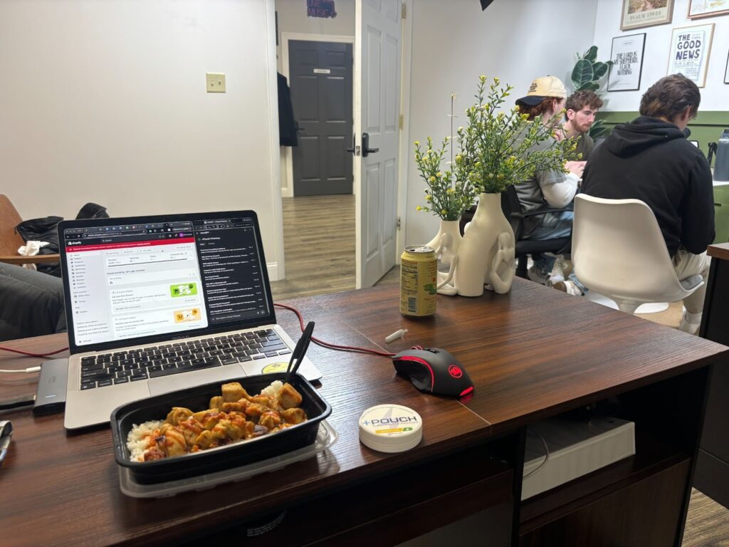 Ryan's laptop sits on a simple, modern desk, with a dish of take out Chinese food in front of it, and a Plus Pouch tin next to it. In the corner two white artsy vases hold yellow evening primose blooms. Behind, three men work at a smaller desk. 