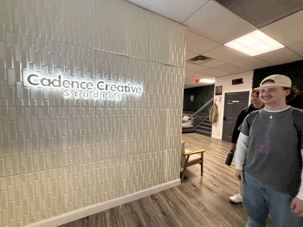 A decorative, textured pattern made to look like a vertical interlocking white brick half wall makes the entry sign backlit to say Cadence Creative Studios. To the right of the sign, we see the beginning of an open floor pattern that shows a lobby chair, a door and coat hooks and a staircase. Plus Pouch CEO stands in front dressed in casual clothes, smiling, with Plus Pouch COO behind him looking toward the Cadence Creative Studios sign. 