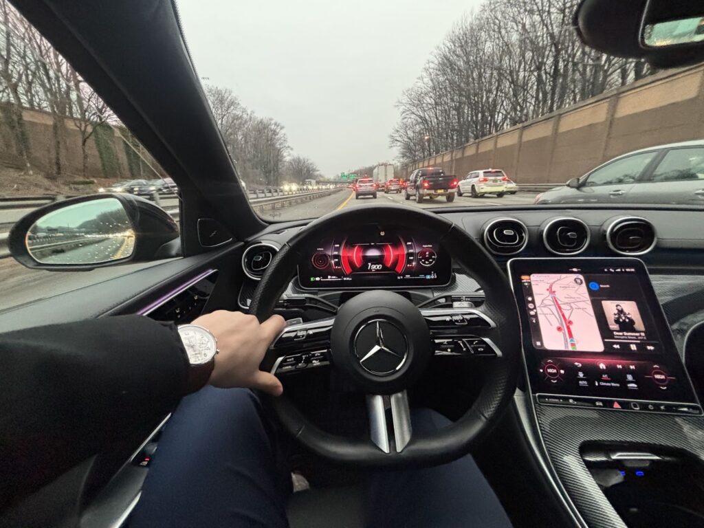 The road ahead of Ryan's car is filled with bright red brake lights on a gray, dreary day, showing standstill traffic on a three-lane highway. Inside the car, we see Ryan's arm and can see he is wearing a suit and a watch. 