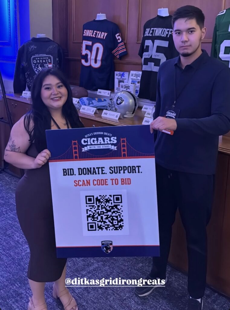 Julia stands with a Rowan sports communication and media student holding QR code signage directing people how to bid on silent auction items at Cigars with the Stars, a pre-Super Bowl event for Gridiron Greats. Behind them we see a table of sports memorabilia. 