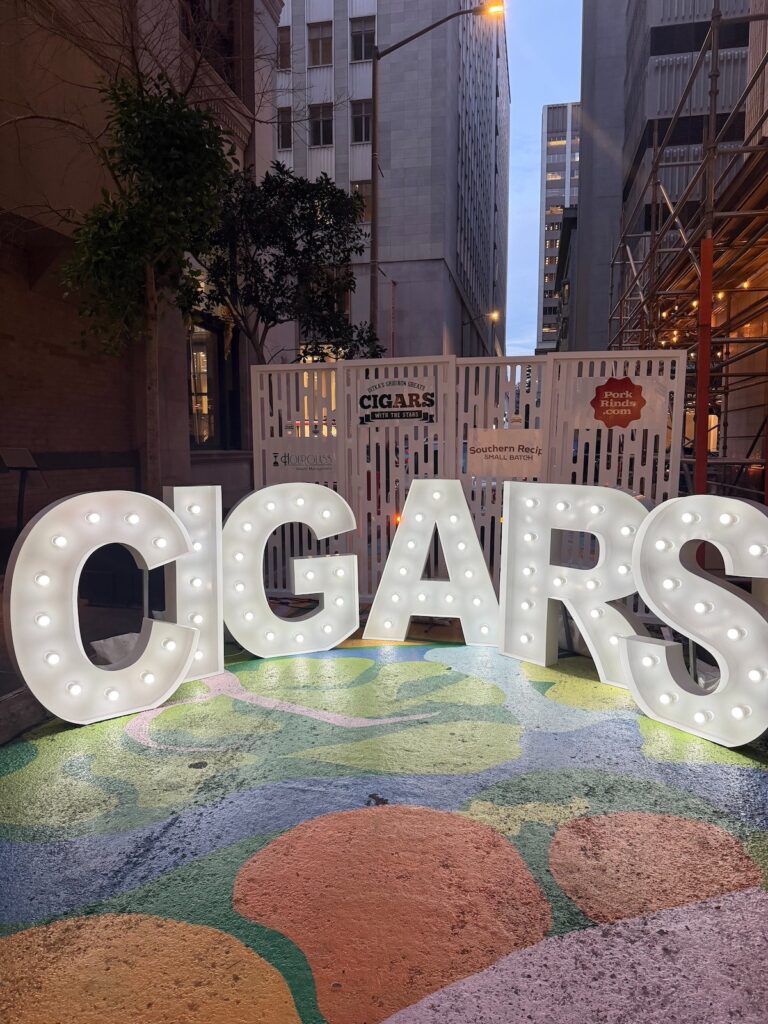 A close up of people-sized light-up letters spelling the word CIGARS, in front of a white entrance gate for the event and the city background of San Francisco in view. 