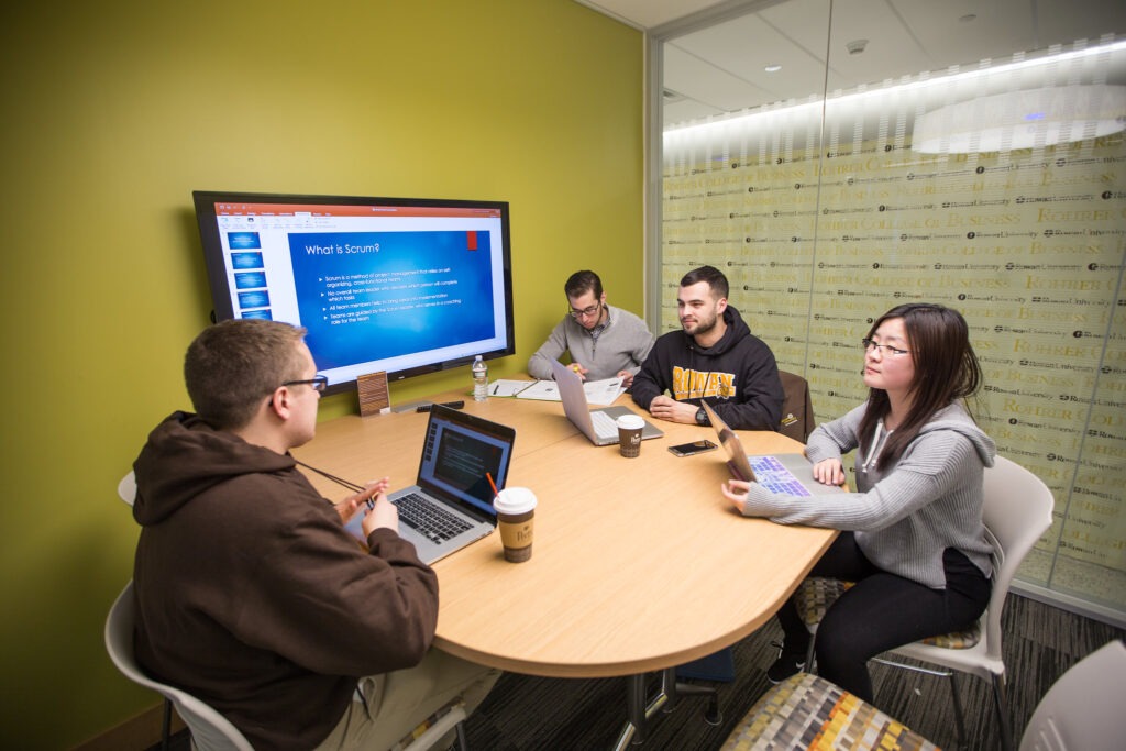 Four college students sitting in a study room with their laptops and other materials with a TV screen that has a slideshow with material.