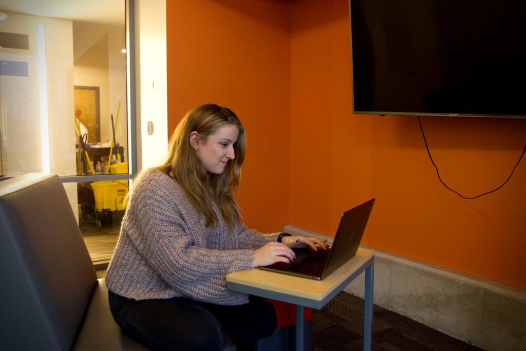College student sitting with laptop open in a study room with TV, preparing for finals.