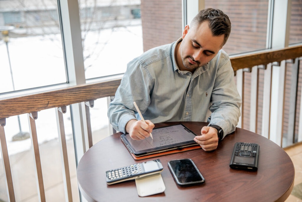 College student sitting at table doing practice problems on his tablet to prepare for final exams.