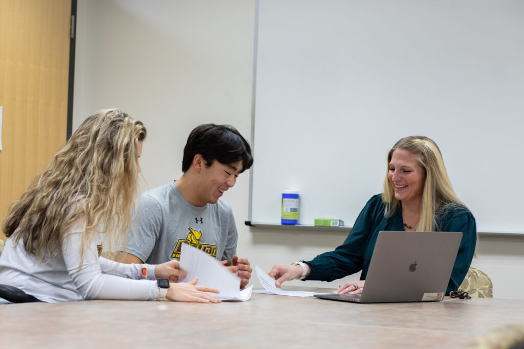 Two college students sitting in a classroom with their professor reviewing materials for their final exam.