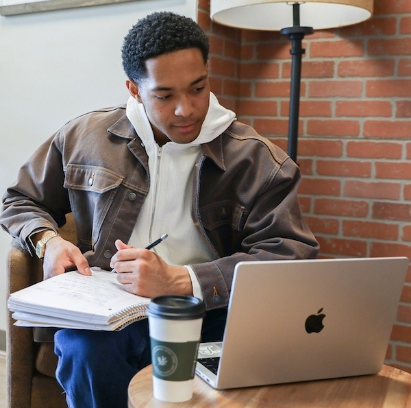 Rowan alumni and MBA candidate, Joshua B., completing coursework in a cafe with laptop open, taking notes.
