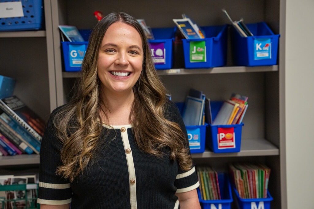 Kaitlyn G., MA in Reading graduate, smiling in Rowan's Reading Clinic in front of blue bookcases.