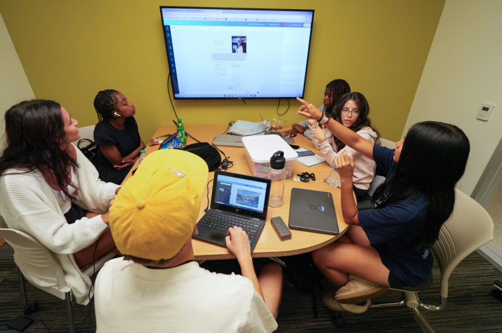 Rowan University students sit around a table with laptops while looking at a shared screen and discussing a digital project.