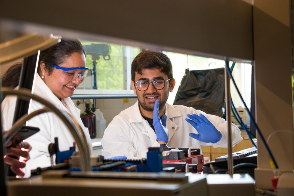 Two college students wearing lab coats, gloves and safety goggles collaborate during a hands-on laboratory internship or research experience.