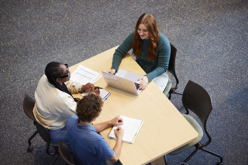 A group of Rowan University students sit around a table with notebooks, folders and a laptop while studying together in a campus building.