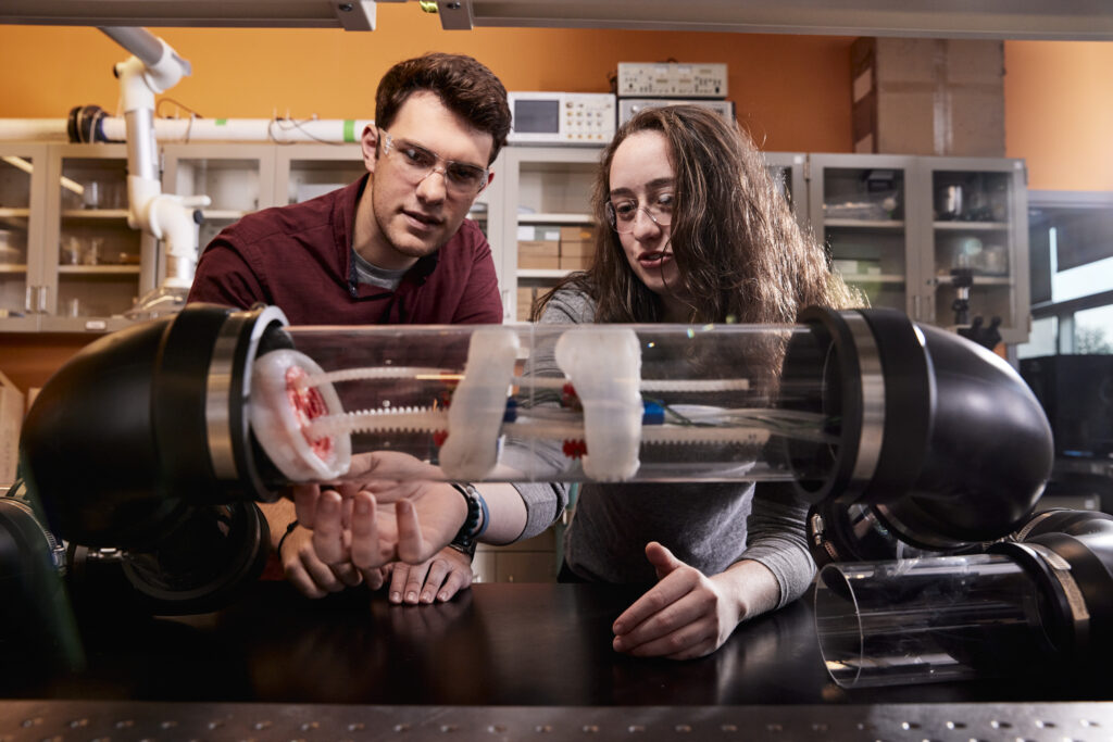 Two Rowan University students wearing safety glasses work together in a lab while examining a clear tube apparatus during a hands-on experiment.