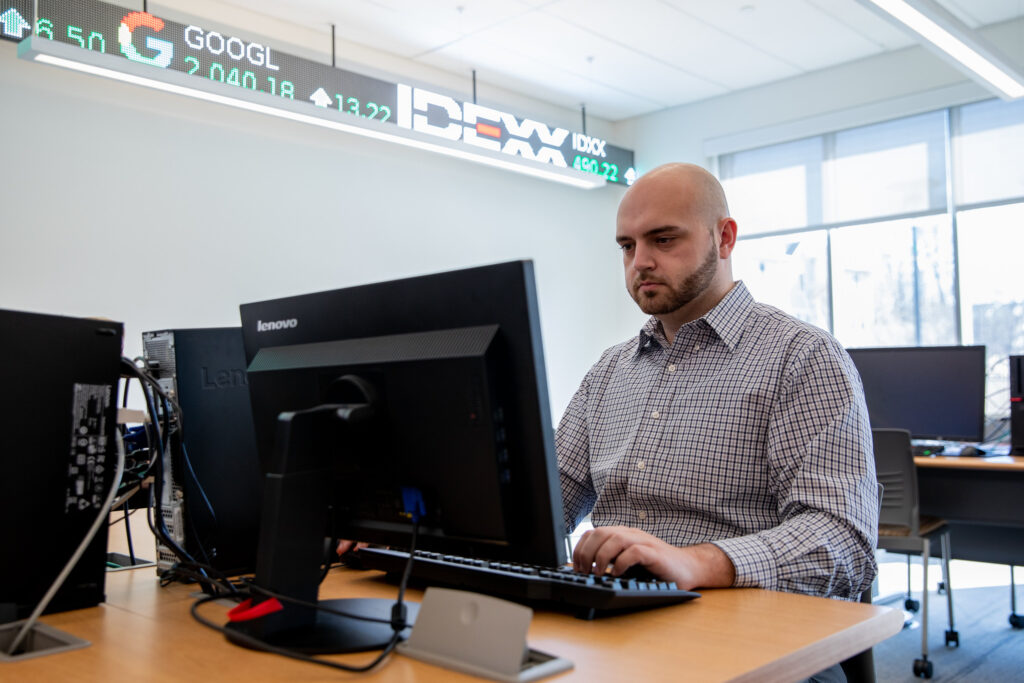 Man working at a desktop computer in a finance and technology classroom with stock ticker displays above the monitors.