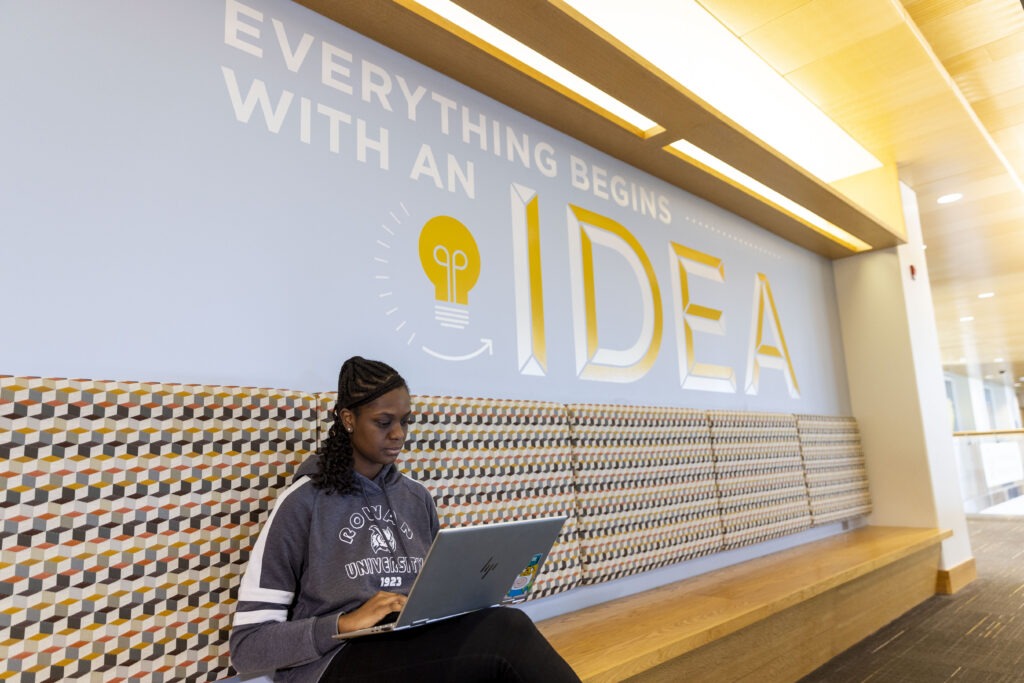 Entrepreneurship student sitting in business hall with laptop in front of an "Everything Begins With an Idea" backdrop.