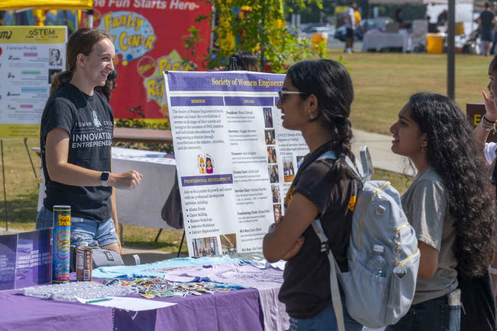 Students speak with a representative at a campus involvement table for the Society of Women Engineers during an outdoor Rowan University event.