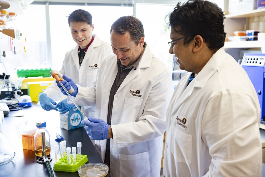 Rowan University students and faculty in white lab coats smile while working together with lab equipment and a pipette during a hands-on research experience.