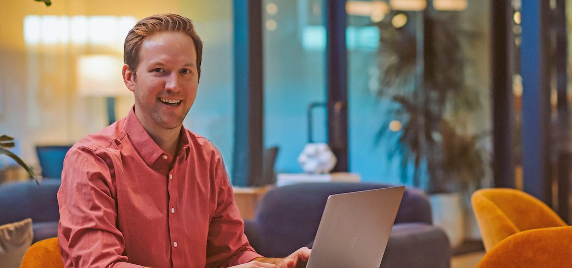Man in a pink button-down shirt smiling while working on a laptop in a modern office lounge area.