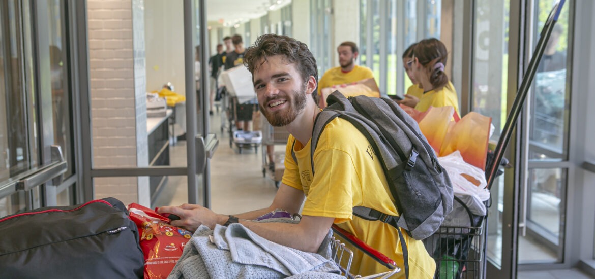 A Rowan University student smiles while pushing a cart filled with dorm supplies through a residence hall during move-in day.