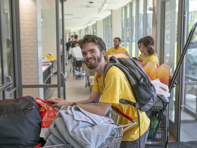 A Rowan University student smiles while pushing a cart filled with dorm supplies through a residence hall during move-in day.
