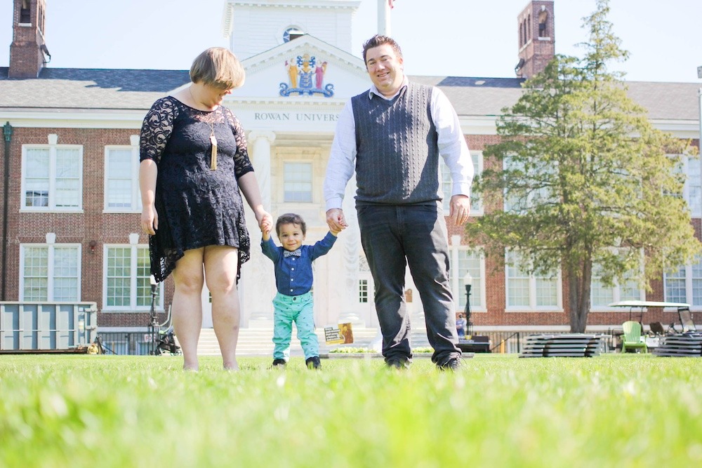 A Rowan University student mother and her husband each hold one hand of their infant who is learning how to walk. The mom is looking down at the child, with the child and husband looking at the camera. The iconic Bunce Hall is behind them. 