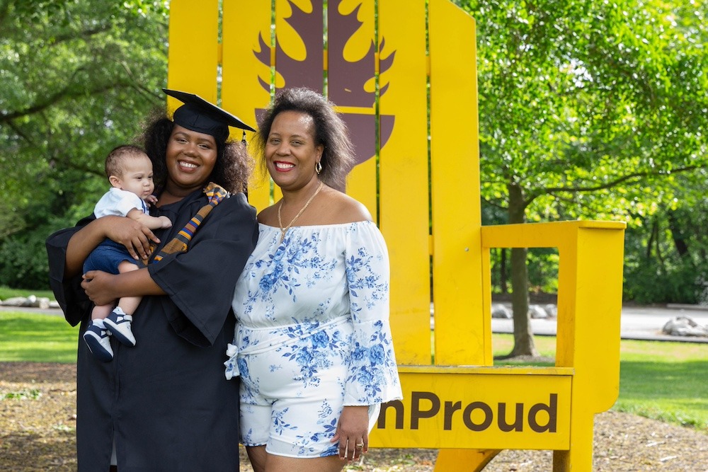 A Rowan University graduate poses for a graduation photo while wearing her cap and gown, standing in front of the oversized yellow #RowanPROUD Adirondak chair, holding her newborn son in her arms, with her mother by her side. 