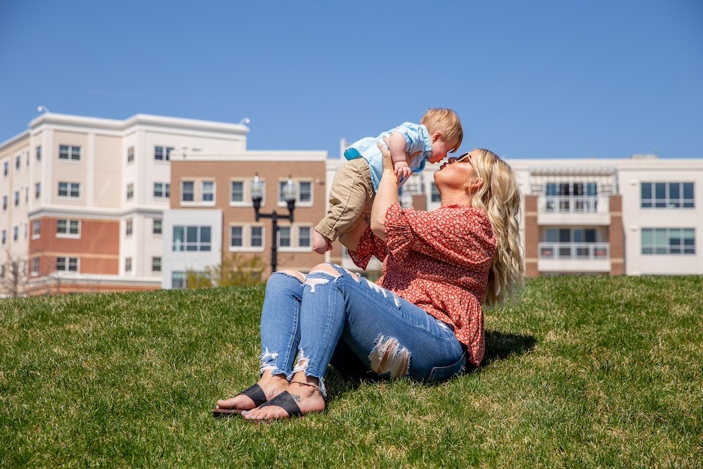 A Rowan University student and mother sits on a a grassy hill on campus, holding her infant slightly above her as she is reaching in to kiss his cute little face. 