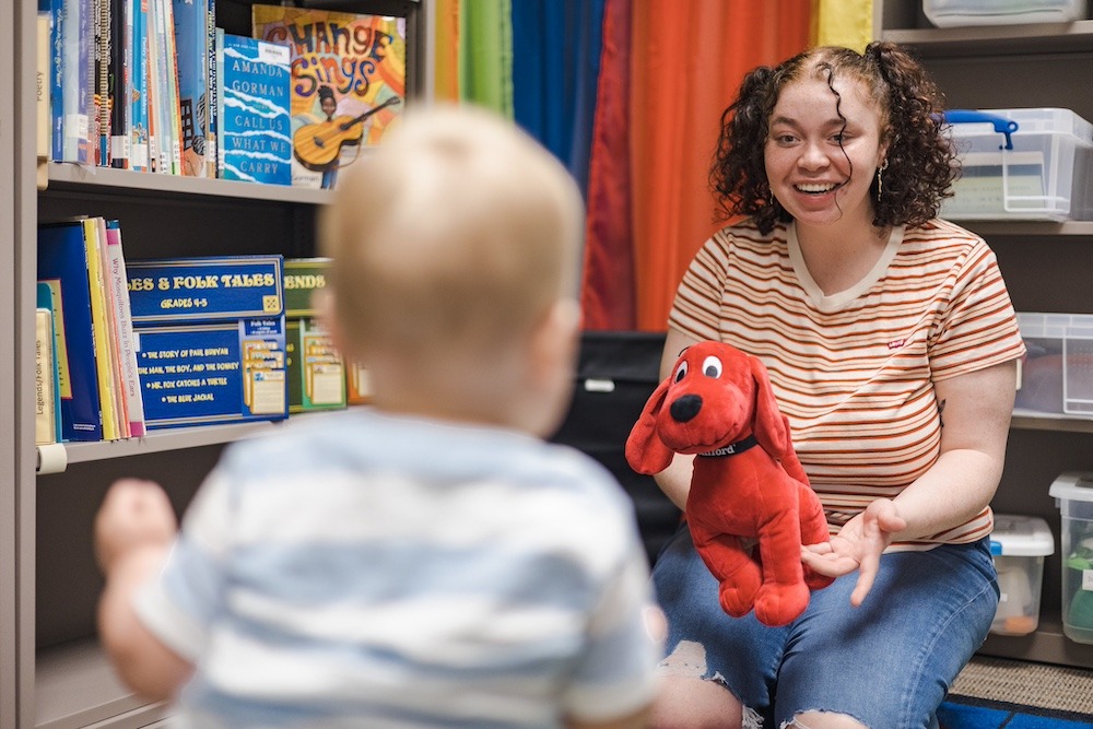 A Rowan University education major holds a stuffed animal of Clifford the Big Red Dog out to a baby who is sitting in front of her.