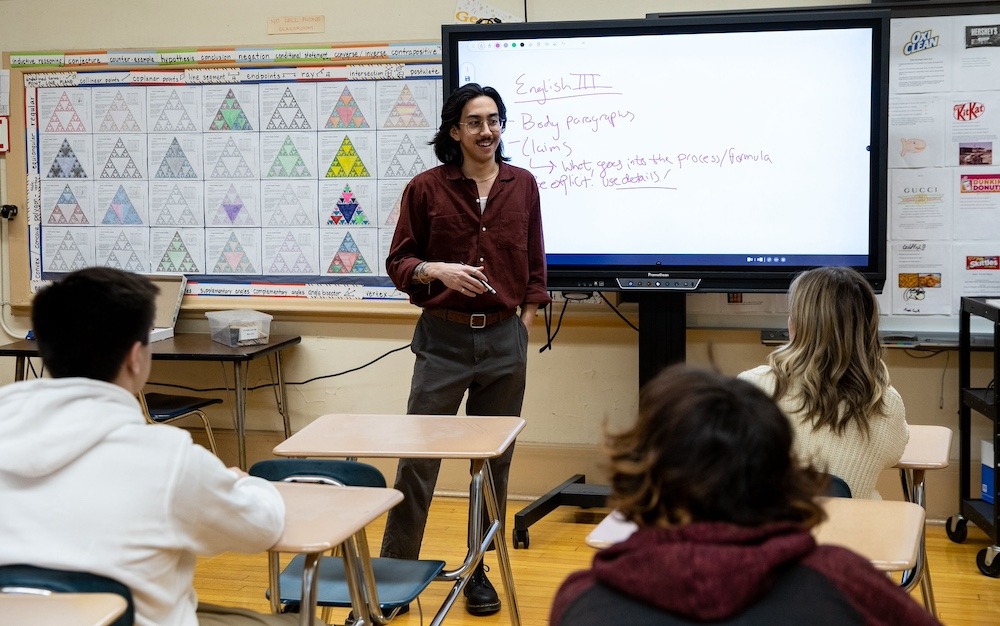 A Rowan University master of science in teaching candidate leads a classroom lesson in a high school, with an electronic whiteboard behind him and seated students in desks in front of him.