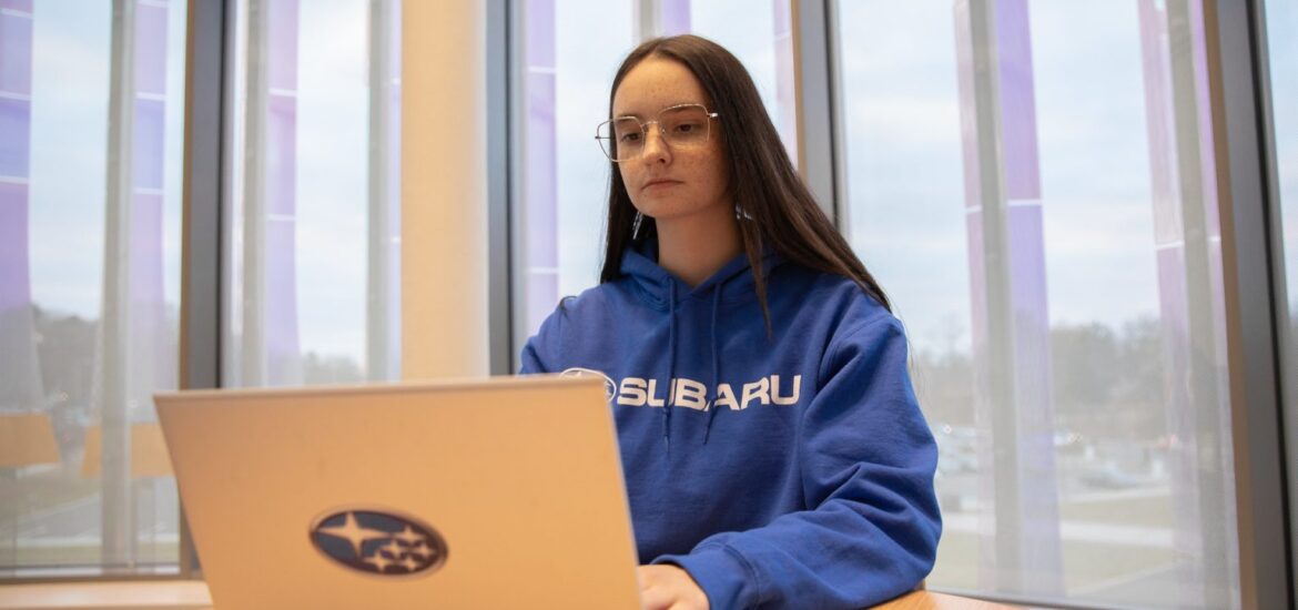 Rowan University Accounting major Jade K. wears a blue sweatshirt and works on a laptop with the Subaru logo in Business Hall.