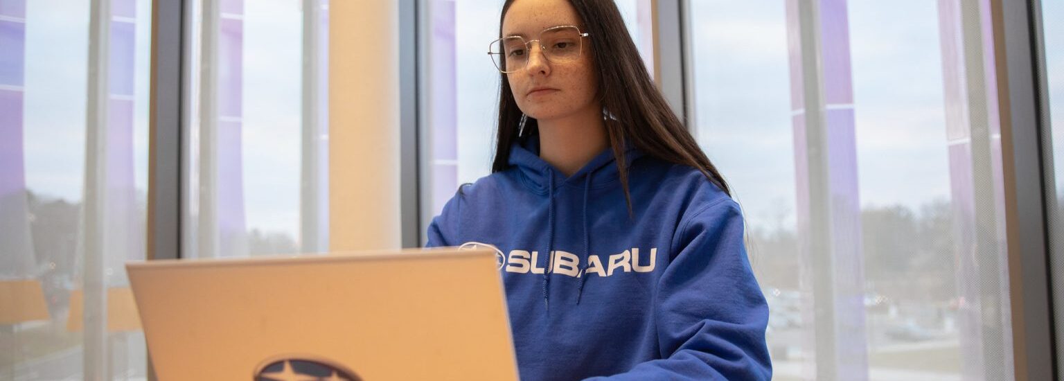 Rowan University Accounting major Jade K. wears a blue sweatshirt and works on a laptop with the Subaru logo in Business Hall.