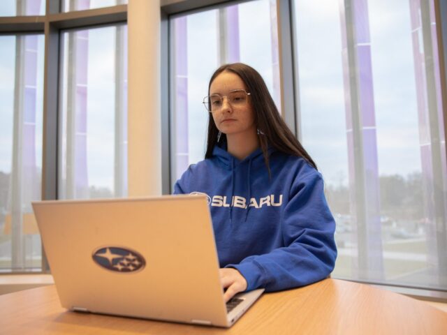 Rowan University Accounting major Jade K. wears a blue sweatshirt and works on a laptop with the Subaru logo in Business Hall.