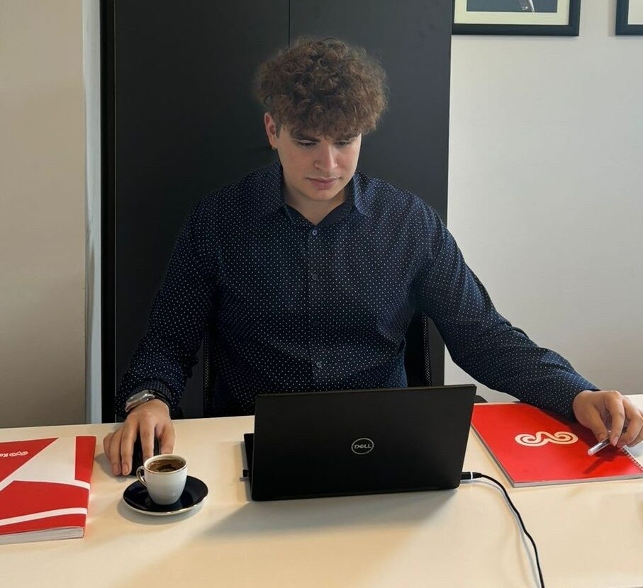Rowan University finance major Tyler G. sits at a desk, flanked by red folders representing the business on either side of his laptop, and a cup of espresso at his fingertips. 