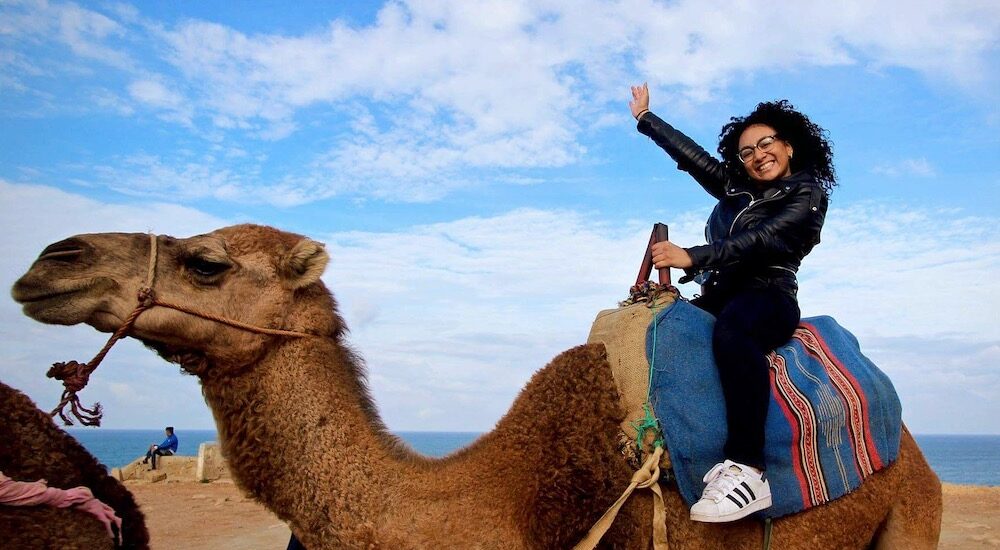 Woman smiling and waving while riding a camel near the ocean under a blue sky.