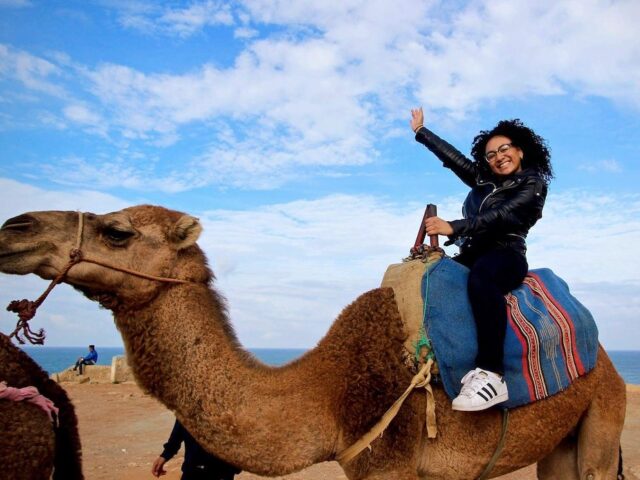 Woman smiling and waving while riding a camel near the ocean under a blue sky.