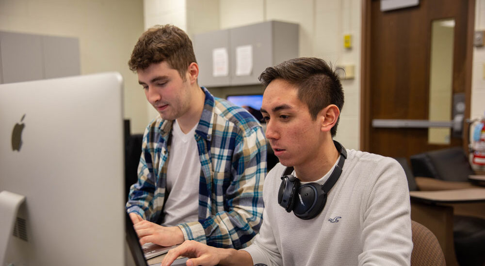 Two male students working together at a desktop computer in a classroom.