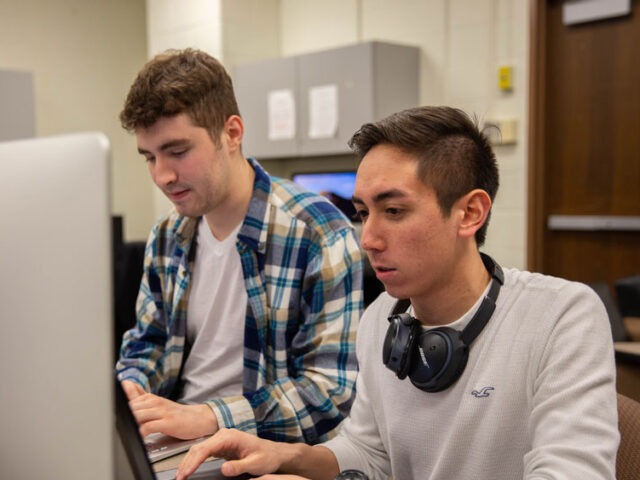 Two male students working together at a desktop computer in a classroom.