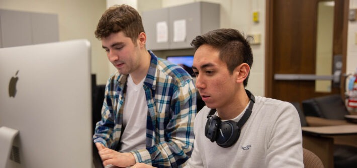 Two male students working together at a desktop computer in a classroom.