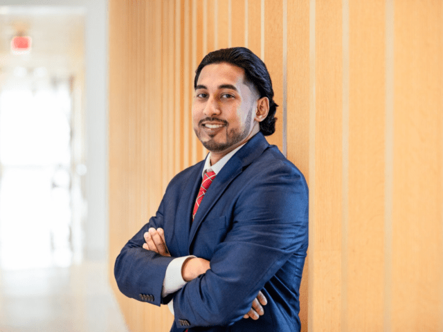 Man in professional business attire standing confidently in a university building hallway.