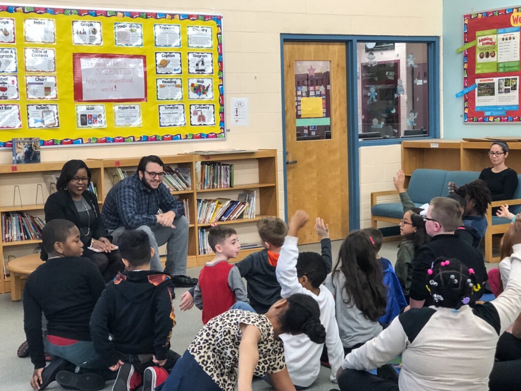 Two educators read to a classroom of elementary school age children who are all sitting on the floor in front of them. 