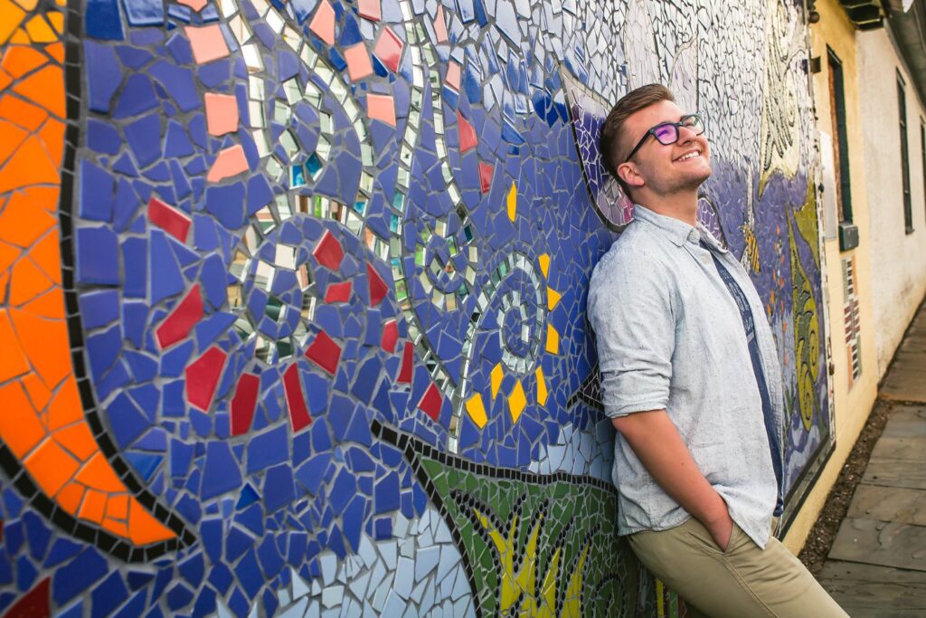 A young educator leans against a school mural made out of mosaic tiles, as he looks slightly upward while smiling. 