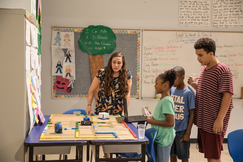 An educator brings a line of students to a table in her classroom to pick up supplies for a project. 