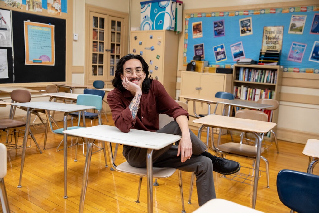 An educator smiles broadly in his empty classroom, sitting at a student desk with his chin resting in his hand as his arm is propped up on the desk. 