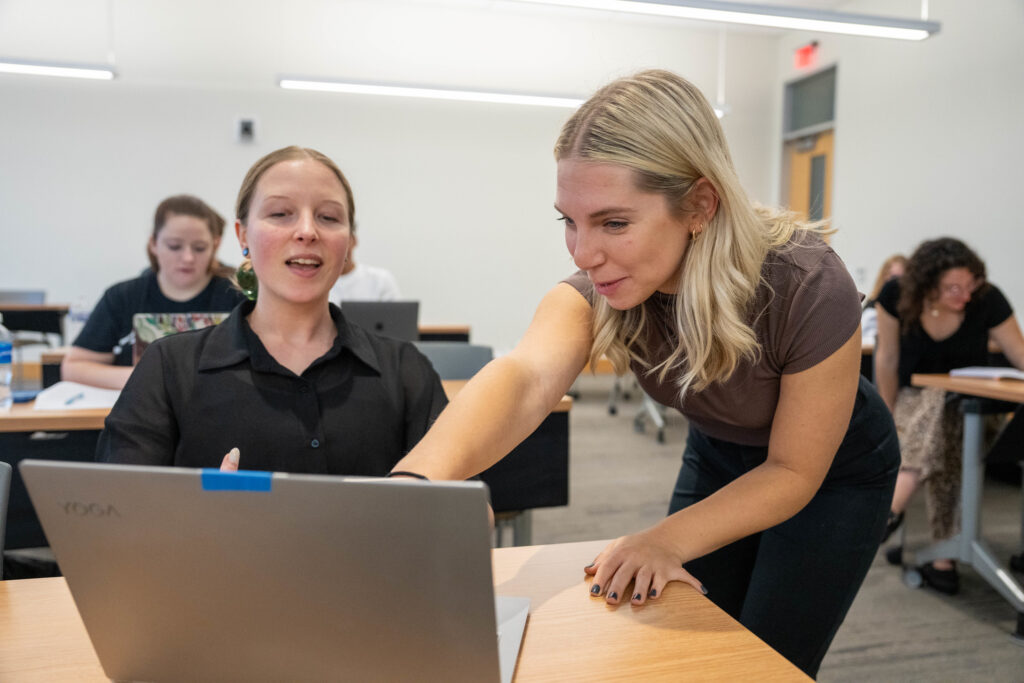 A teacher leans down in a classroom to point to a student's laptop. 